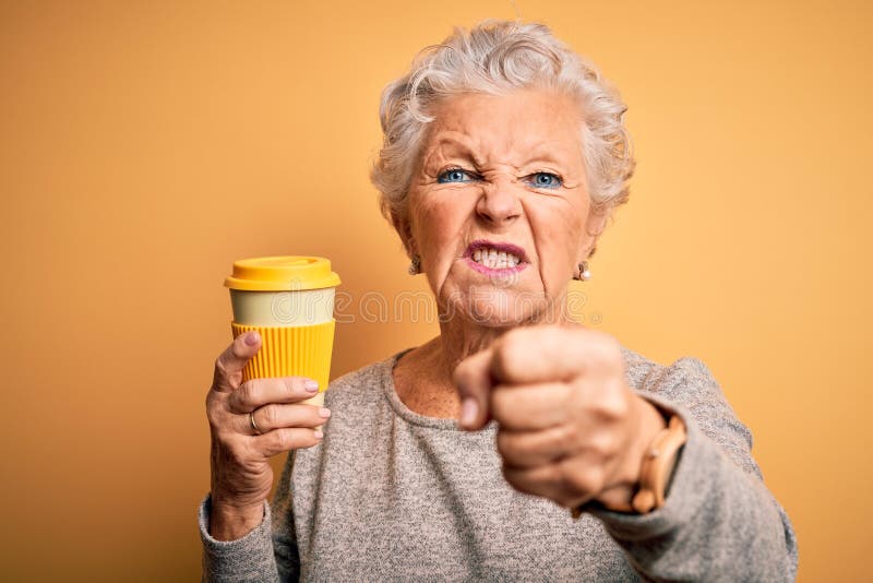 Högre, vacker kvinna dricker kaffe som står över isolerad gul bakgrund irriterad och frustrerad skrik med ilska. En vacker kvinnlig kopp kaffe som står över isolerad gul bakgrund irriterad och frustrerad skrik med ilska och skrik med ett upphöjt "hand iland"-koncept