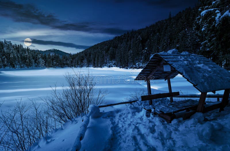 Wooden bower in snowy winter spruce forest. beautiful mountainous landscape near snow covered frozen lake at night in full moon light. Wooden bower in snowy winter spruce forest. beautiful mountainous landscape near snow covered frozen lake at night in full moon light