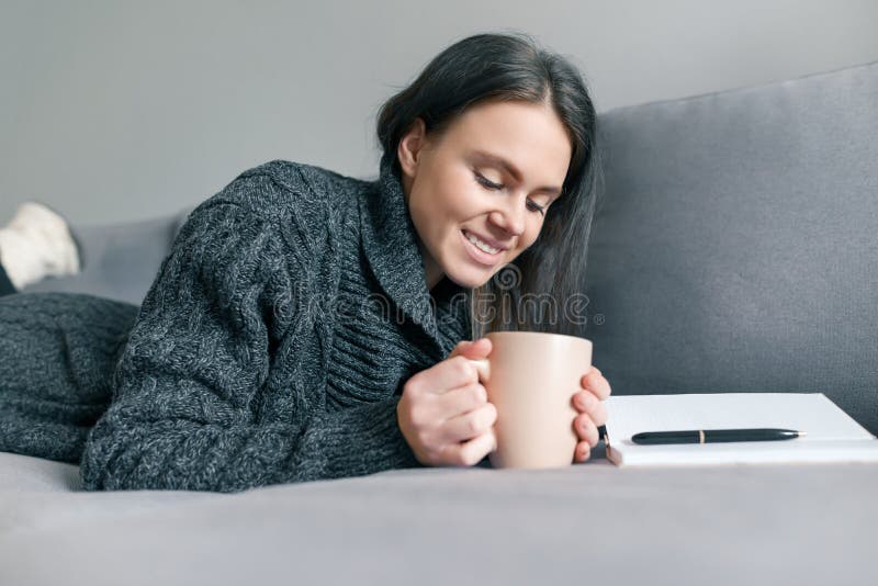 Höstvinterstående av unga flickan i varm stucken tröja hemma på soffan med anteckningsboken och koppen av den varma drinken royaltyfri foto Höstvinterstående av unga flickan i varm stucken tröja hemma på soffan med anteckningsboken och koppen av den varma drinken royaltyfri foto