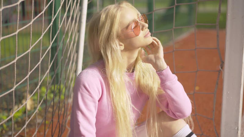 Hübsche blonde Frau in der Sonnenbrillen, die an den Fußballtoren am sonnigen Sommertag sitzt. Porträt des jungen reizend Kaukasie stock video footage