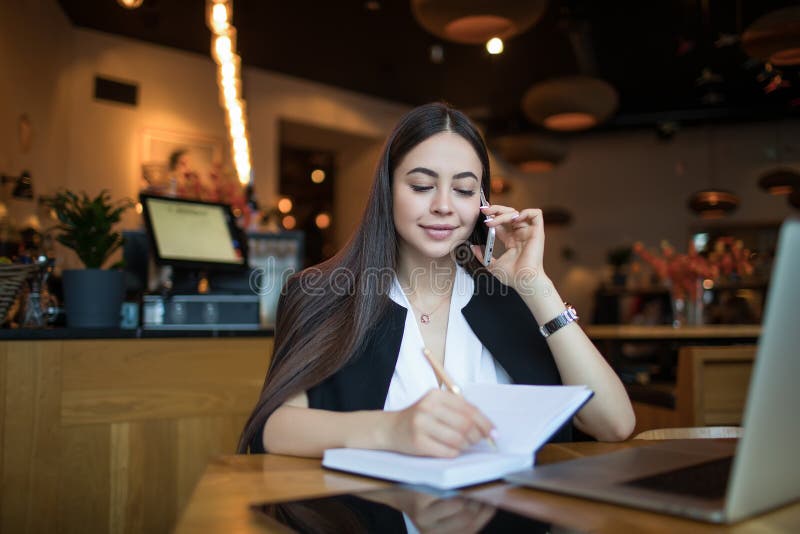 Hübsche internationale Studentin, die Informationen in Textbüchern schreiben und über Smartphone sprechen lizenzfreie stockbilder