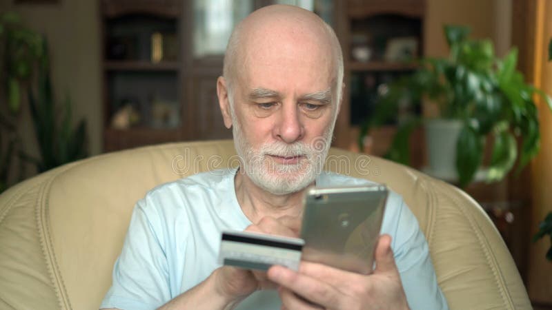 Hübscher lächelnder älterer Mann, der zu Hause auf Stuhl sitzt Online kaufen mit Kreditkarte auf Smartphone