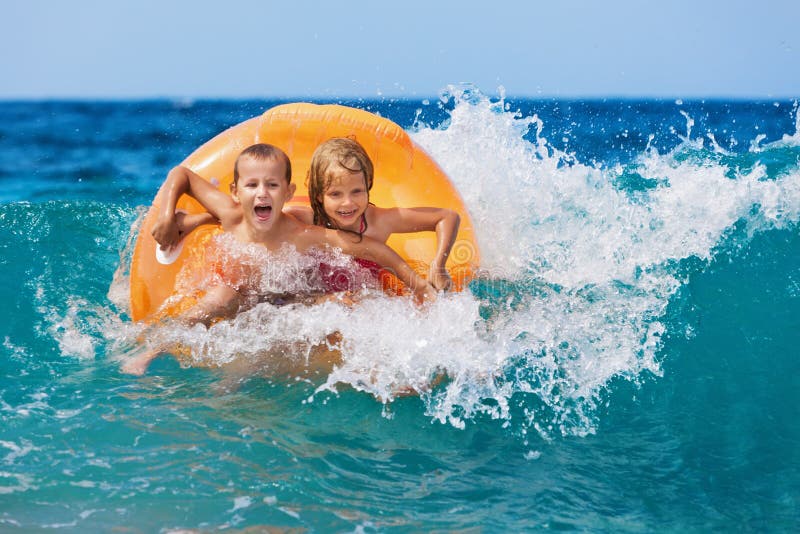 Happy kids have fun in sea surf on beach. Joyful couple of children on inflatable ring ride on breaking wave. Travel lifestyle, swimming activities in family summer camp. Vacations on tropical island. Happy kids have fun in sea surf on beach. Joyful couple of children on inflatable ring ride on breaking wave. Travel lifestyle, swimming activities in family summer camp. Vacations on tropical island