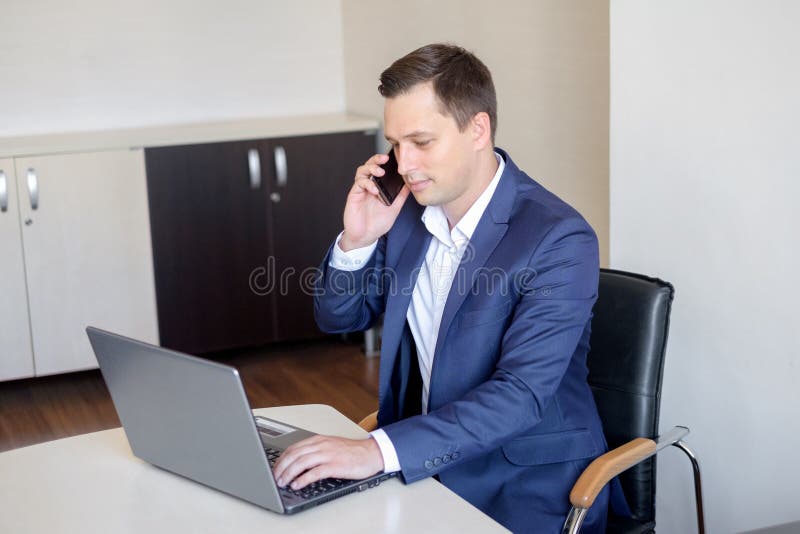 I giovani bei hanno concentrato l'uomo attraente che utilizza il cellulare mentre lavoravano con il computer portatile nell'uffic fotografia stock libera da diritti I giovani bei hanno concentrato l'uomo attraente che utilizza il cellulare mentre lavoravano con il computer portatile nell'uffic fotografia stock libera da diritti