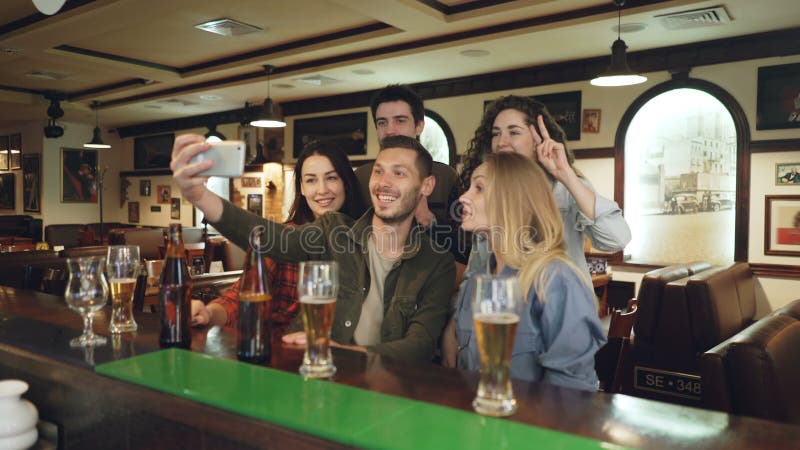 I giovani stanno utilizzando lo smartphone per prendere il selfie nella barra popolare Gli amici sono posanti, divertentesi, gest