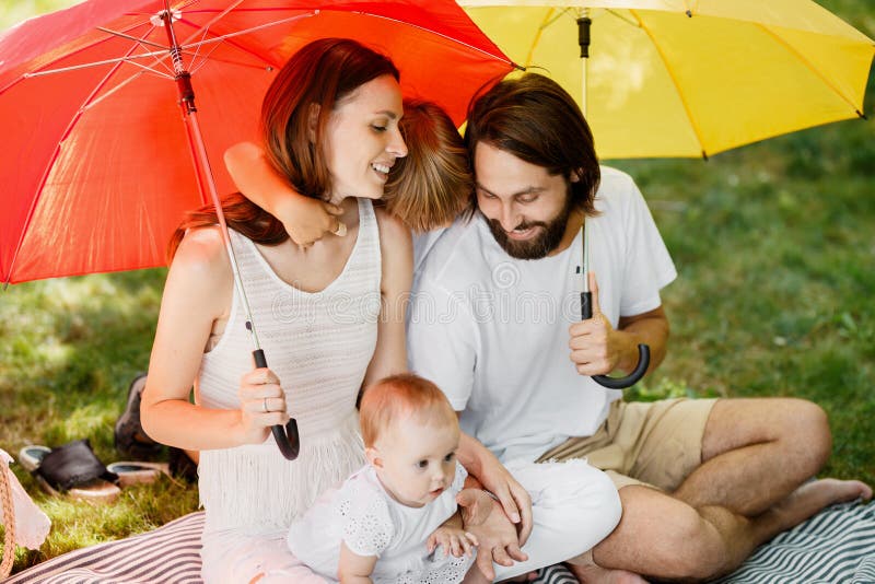 I grandi ombrelli luminosi coprono la famiglia felice vestita nei vestiti bianchi che si siedono sulla coperta dal sole fotografia stock I grandi ombrelli luminosi coprono la famiglia felice vestita nei vestiti bianchi che si siedono sulla coperta dal sole fotografia stock