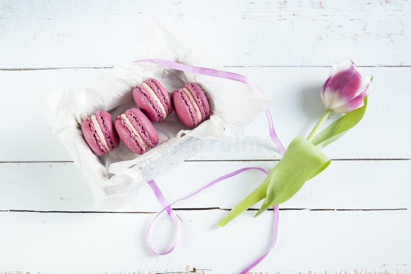 I maccheroni francesi cremisi dolci con la scatola ed il tulipano su luce hanno tinto il fondo di legno immagini stock libere da diritti I maccheroni francesi cremisi dolci con la scatola ed il tulipano su luce hanno tinto il fondo di legno immagini stock libere da diritti