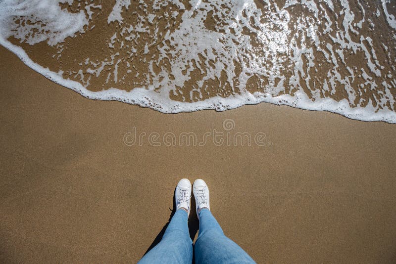 I piedi della donna osservano con i jeans da sopra alla spiaggia con la sabbia e l'onda che viene nel telaio immagini stock libere da diritti I piedi della donna osservano con i jeans da sopra alla spiaggia con la sabbia e l'onda che viene nel telaio immagini stock libere da diritti