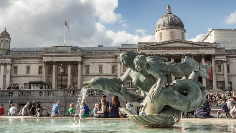 I turisti si siedono dalla fontana in Trafalgar Square con la statua ed il National Gallery della sirena fotografie stock libere da diritti I turisti si siedono dalla fontana in Trafalgar Square con la statua ed il National Gallery della sirena fotografie stock libere da diritti