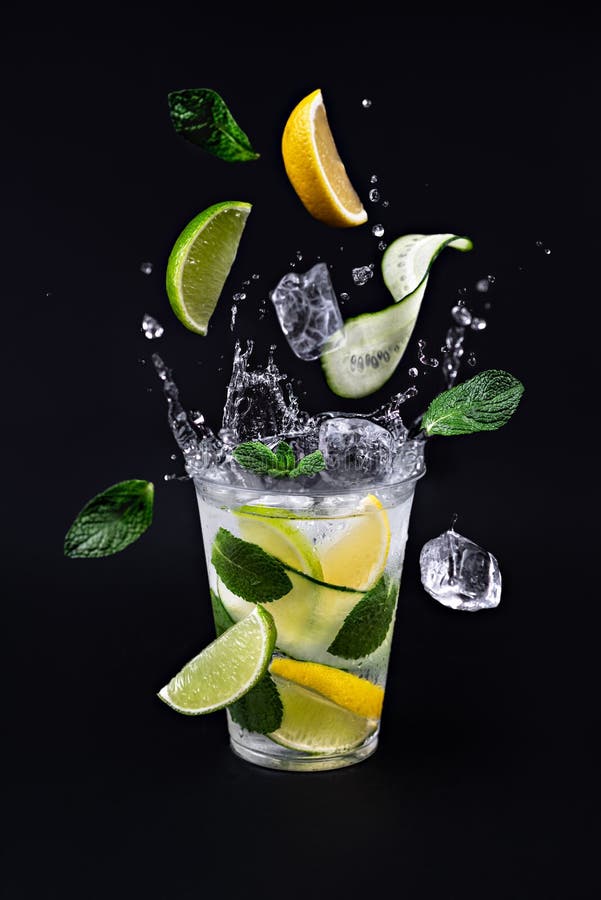 Ice cubes, slices of lime. lemon, cucumber and mint leaves splashing into glass of lemonade on black