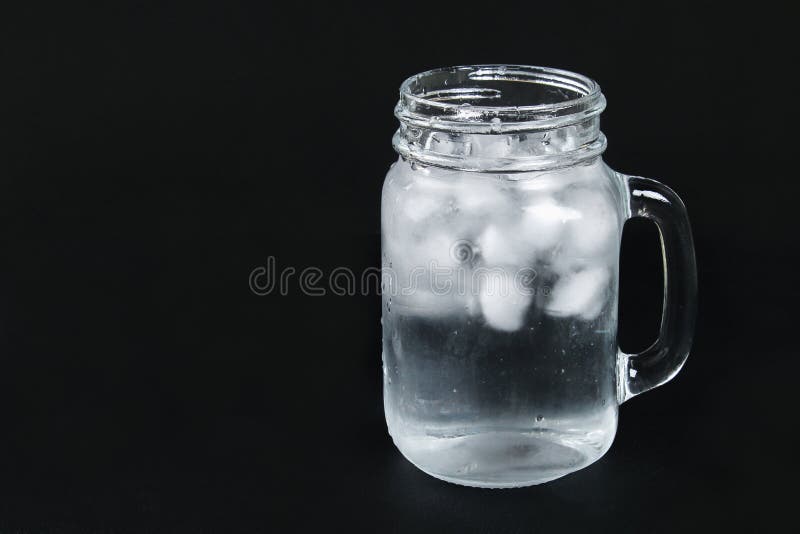 Ice water with ice and a mint leaf and a straw in a mason jar on a black background.