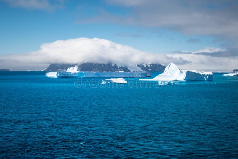 Travel image with winter landscape in Antarctica. Seascape with Icebergs in the Antarctic Ocean, mountain covered with big cloud in the background. Travel image with winter landscape in Antarctica. Seascape with Icebergs in the Antarctic Ocean, mountain covered with big cloud in the background.