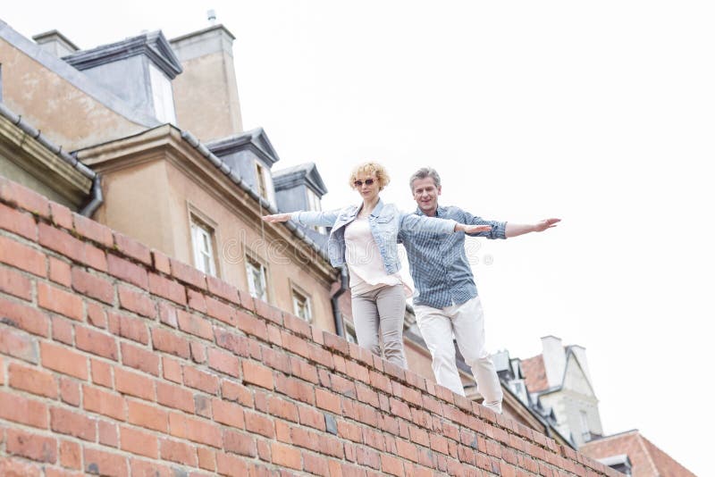 Low angle view of middle-aged couple with arms outstretched walking on brick wall against clear sky. Low angle view of middle-aged couple with arms outstretched walking on brick wall against clear sky