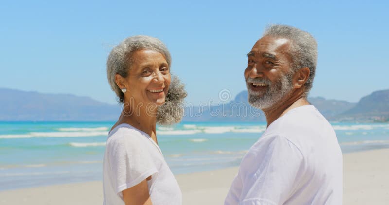 Ideia lateral dos pares afro-americanos superiores ativos felizes que estão na praia na luz do sol 4k