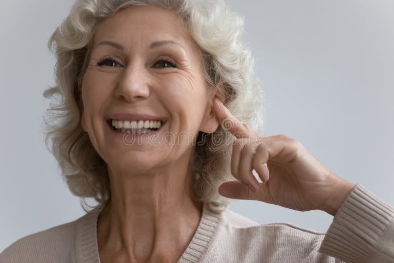 Idosa aponta o dedo ao ouvido com aparelho de assistência auditiva