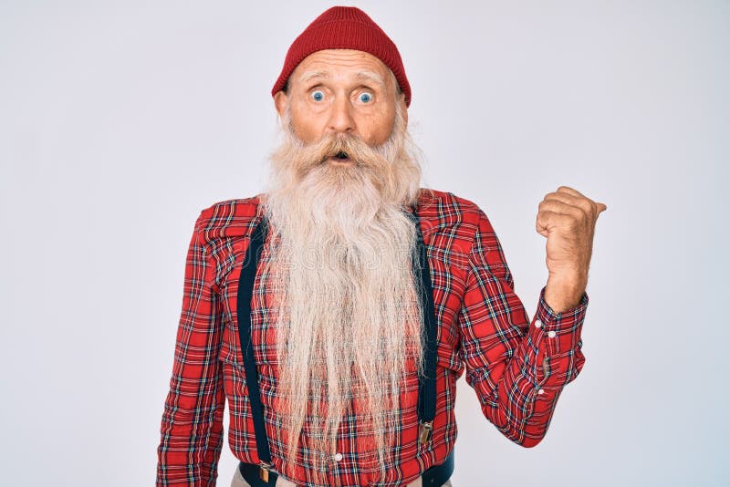 Old senior man with grey hair and long beard wearing hipster look with wool cap surprised pointing with hand finger to the side, open mouth amazed expression. Old senior man with grey hair and long beard wearing hipster look with wool cap surprised pointing with hand finger to the side, open mouth amazed expression