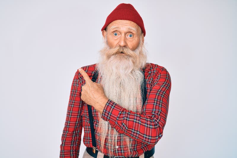 Old senior man with grey hair and long beard wearing hipster look with wool cap surprised pointing with finger to the side, open mouth amazed expression. Old senior man with grey hair and long beard wearing hipster look with wool cap surprised pointing with finger to the side, open mouth amazed expression