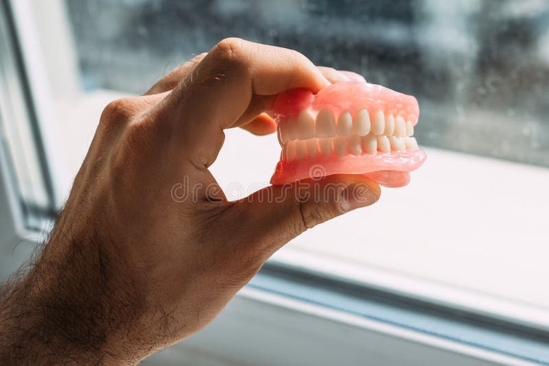 An elderly man holds a denture. A man is holding dentures in his hands. Removable dentures flexible. False teeth. Implant. Orthodontics. Old age. Teeth. Jaw. The close plan. Removable denture. An elderly man holds a denture. A man is holding dentures in his hands. Removable dentures flexible. False teeth. Implant. Orthodontics. Old age. Teeth. Jaw. The close plan. Removable denture