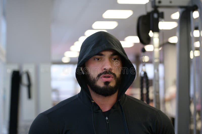 Idrottsmotivation: ung skäggig stark man i hoodie i träningshym med sinneskoncentration royaltyfri bild
