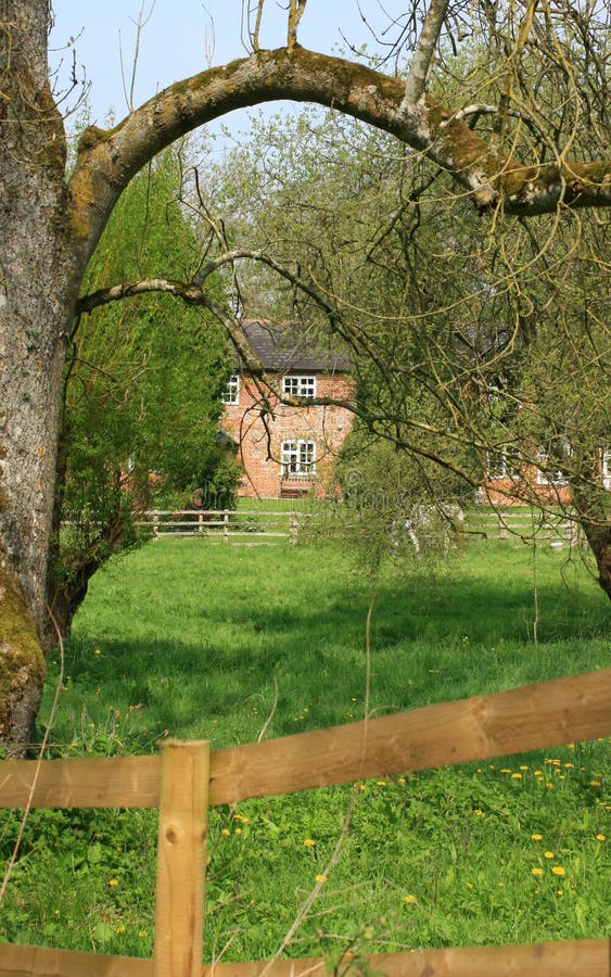 A red bricked country cottage in rural Wiltshire, England viewed across a green grassed field and trees. A paddock fence to foreground. A red bricked country cottage in rural Wiltshire, England viewed across a green grassed field and trees. A paddock fence to foreground.