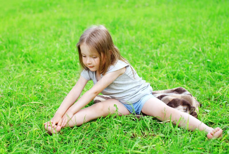 Il bambino della bambina che si siede sull'erba fa l'allungamento di yoga immagine stock Il bambino della bambina che si siede sull'erba fa l'allungamento di yoga immagine stock