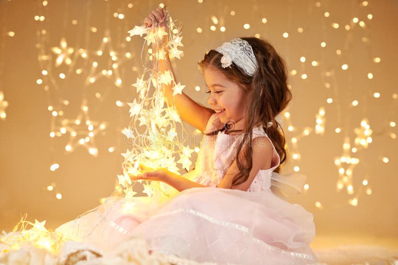 Il bambino della ragazza sta giocando con le luci di natale, il fondo giallo, vestito rosa fotografia stock Il bambino della ragazza sta giocando con le luci di natale, il fondo giallo, vestito rosa fotografia stock