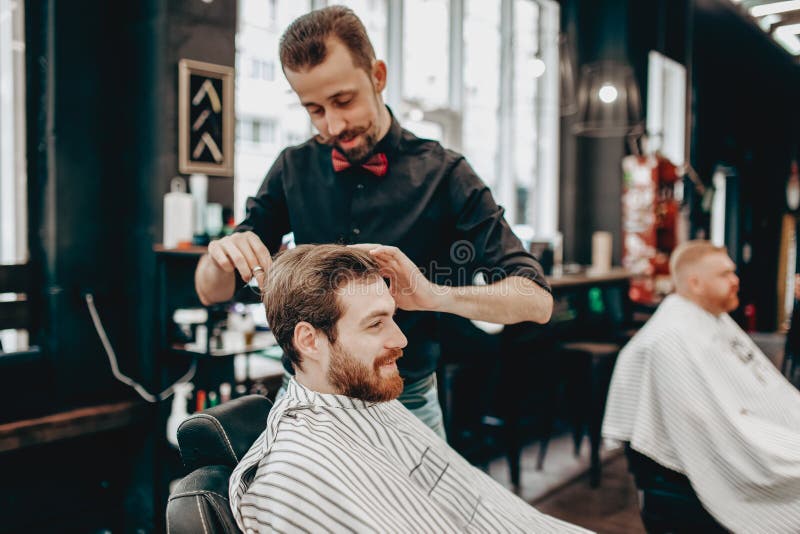 Il barbiere baffuto vestito in una camicia nera con una cravatta a farfalla rossa fa un'acconciatura alla moda al giovane in un p immagine stock libera da diritti Il barbiere baffuto vestito in una camicia nera con una cravatta a farfalla rossa fa un'acconciatura alla moda al giovane in un p immagine stock libera da diritti