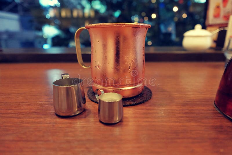 Il caffè nero ghiacciato contenuto in una tazza di rame è servito con sciroppo e latte fresco con la scena confusa di notte fotografie stock Il caffè nero ghiacciato contenuto in una tazza di rame è servito con sciroppo e latte fresco con la scena confusa di notte fotografie stock