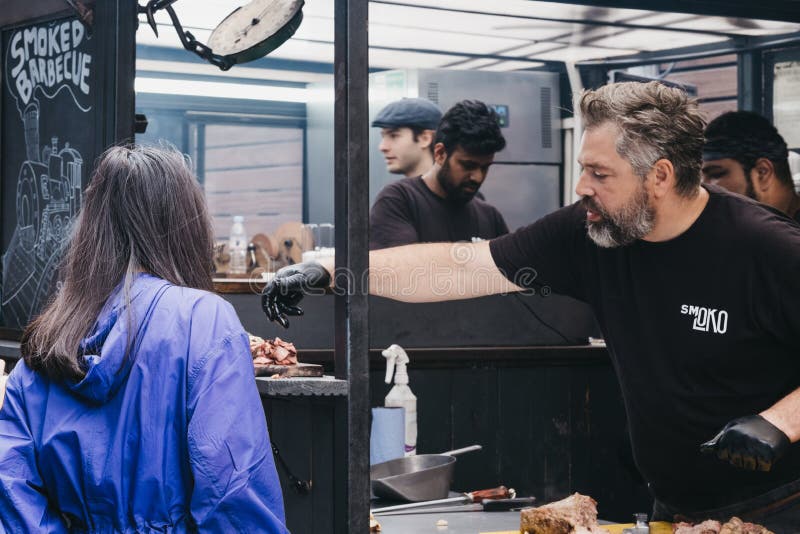 Il campionamento della donna ha grigliato la carne ad una stalla di Smokoloco dentro il mercato di Spitalfields, Londra, Regno Un fotografie stock Il campionamento della donna ha grigliato la carne ad una stalla di Smokoloco dentro il mercato di Spitalfields, Londra, Regno Un fotografie stock