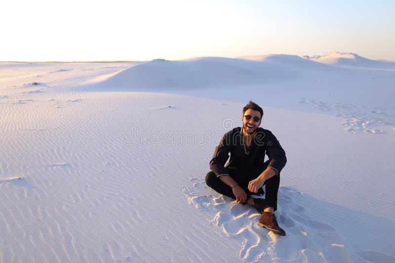 Handsome Emirate man sits on white sand of desert, possing and smiles broad smile, looks around neighborhood on clear evening. Swarthy Muslim with short dark hair and sunglasses dressed in black kandur, long spacious dress made of dark cotton and dark brown shoes are tied. Concept of Arab and Muslim men, united Arab emirates and beautiful landscapes, advertising services of travel companies or airlines, national clothes of Emirates, good mood and enjoyment of life. Handsome Emirate man sits on white sand of desert, possing and smiles broad smile, looks around neighborhood on clear evening. Swarthy Muslim with short dark hair and sunglasses dressed in black kandur, long spacious dress made of dark cotton and dark brown shoes are tied. Concept of Arab and Muslim men, united Arab emirates and beautiful landscapes, advertising services of travel companies or airlines, national clothes of Emirates, good mood and enjoyment of life.
