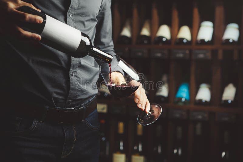 Il colpo alto vicino del sommelier che versa il vino rosso da imbottiglia il vetro sul fondo sotterraneo della cantina fotografia stock Il colpo alto vicino del sommelier che versa il vino rosso da imbottiglia il vetro sul fondo sotterraneo della cantina fotografia stock