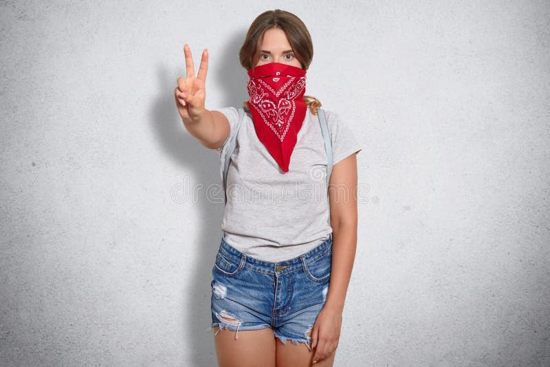 Indoor shot of self confident female feminist shows peace sign, wears fashionable red bandana, wears jean shorts and grey t shirt, isolated over grey background. All woman are equal with men. Indoor shot of self confident female feminist shows peace sign, wears fashionable red bandana, wears jean shorts and grey t shirt, isolated over grey background. All woman are equal with men