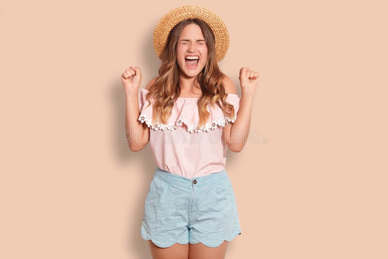 Il colpo orizzontale di giovane femmina europea allegra serra i pugni, esclama con felicità, chiude gli occhi, porta il cappello fotografia stock Il colpo orizzontale di giovane femmina europea allegra serra i pugni, esclama con felicità, chiude gli occhi, porta il cappello fotografia stock