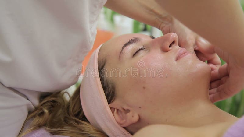 Il cosmetologo femminile che massaggia il fronte di giovane cliente biondo in salone di lusso