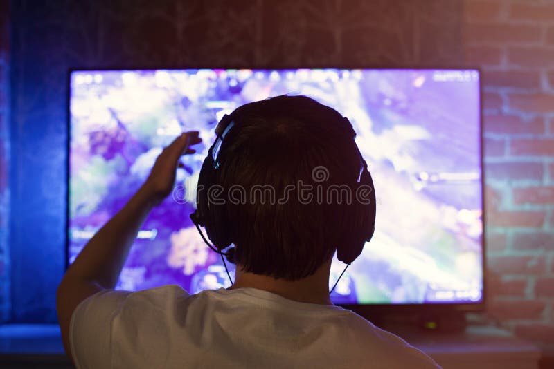 Il Gamer o la fiamma in cuffie con il microfono si siede a casa nella stanza scura e nei giochi con gli amici sulle reti in video immagine stock libera da diritti Il Gamer o la fiamma in cuffie con il microfono si siede a casa nella stanza scura e nei giochi con gli amici sulle reti in video immagine stock libera da diritti