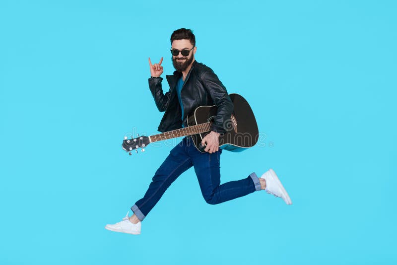Il giocatore di chitarra dell'uomo salta mentre mostra il gesto della roccia fotografia stock Il giocatore di chitarra dell'uomo salta mentre mostra il gesto della roccia fotografia stock