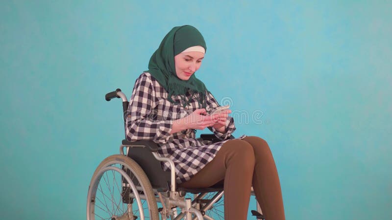 Il giovane disabile musulmano della donna in una sedia a rotelle utilizza il telefono su un fondo blu