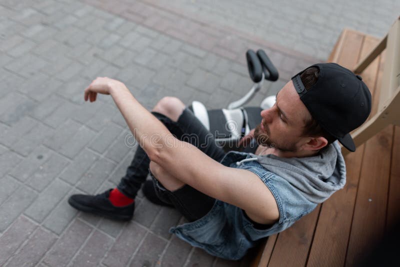 Il giovane moderno americano in un cappuccio d'avanguardia in una maglia del denim in jeans strappati neri alla moda in scarpe da fotografie stock Il giovane moderno americano in un cappuccio d'avanguardia in una maglia del denim in jeans strappati neri alla moda in scarpe da fotografie stock