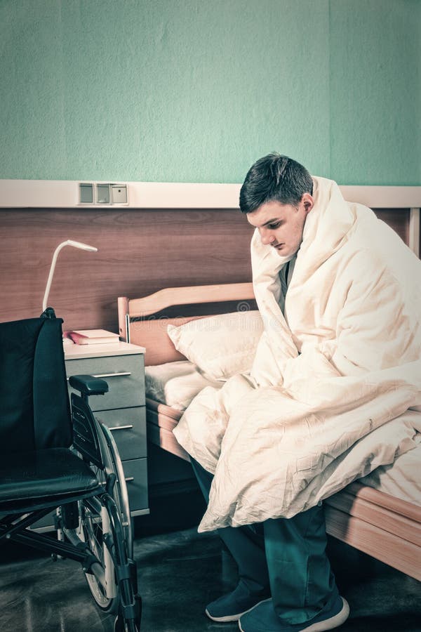 Il giovane paziente malato maschio triste che si siede sul letto di ospedale ha riguardato w immagini stock libere da diritti Il giovane paziente malato maschio triste che si siede sul letto di ospedale ha riguardato w immagini stock libere da diritti