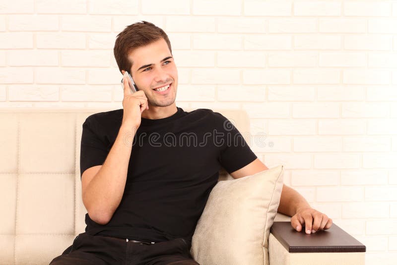 Il giovane uomo attraente in camicia nera sta sedendosi sul sofà e sta parlando sul telefono a casa Copi lo spazio fotografie stock Il giovane uomo attraente in camicia nera sta sedendosi sul sofà e sta parlando sul telefono a casa Copi lo spazio fotografie stock