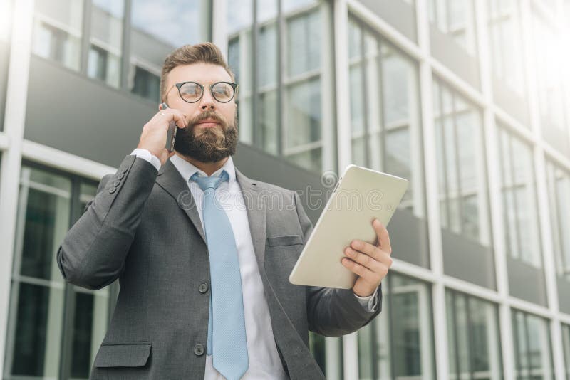 Il giovane uomo d'affari in vestito ed in legame sta stando all'aperto, sta tenendo il computer della compressa e sta parlando su fotografie stock libere da diritti Il giovane uomo d'affari in vestito ed in legame sta stando all'aperto, sta tenendo il computer della compressa e sta parlando su fotografie stock libere da diritti