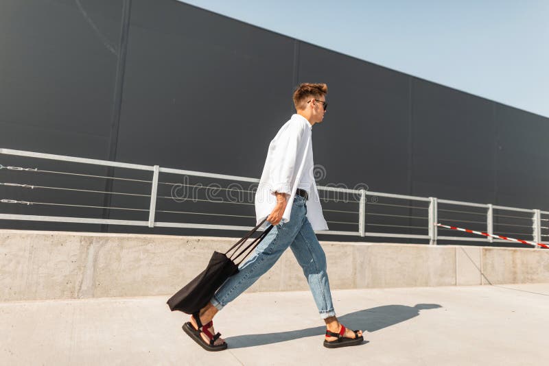 Il giovane uomo europeo dei pantaloni a vita bassa in una camicia bianca in blue jeans in sandali alla moda con una borsa nera ca fotografia stock libera da diritti Il giovane uomo europeo dei pantaloni a vita bassa in una camicia bianca in blue jeans in sandali alla moda con una borsa nera ca fotografia stock libera da diritti