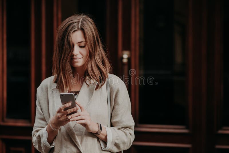 Il giovanotto femminile contentissimo piacevole guarda il video sullo Smart Phone, legge la notifica di attività bancarie, vestit fotografia stock
