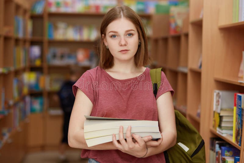 Il giovanotto femminile intelligente sveglio tiene il libro, porta lo Zaino, posa nella biblioteca di scuola, cerca materiale nec fotografia stock libera da diritti