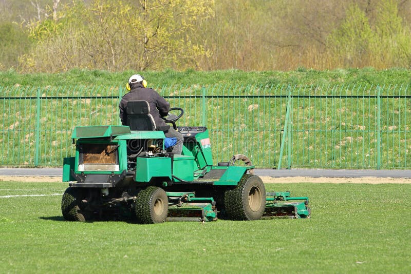 Il lavoratore su una grande falciatrice da giardino verde falcia l'erba sul campo di football americano Architettura del p?saggio immagini stock libere da diritti Il lavoratore su una grande falciatrice da giardino verde falcia l'erba sul campo di football americano Architettura del p?saggio immagini stock libere da diritti