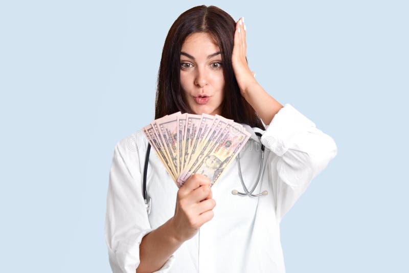 Il medico femminile sgomento con capelli scuri, sguardi sorprendente ai dollari, riceve bribary enorme, implicato nella corruzion fotografia stock libera da diritti Il medico femminile sgomento con capelli scuri, sguardi sorprendente ai dollari, riceve bribary enorme, implicato nella corruzion fotografia stock libera da diritti