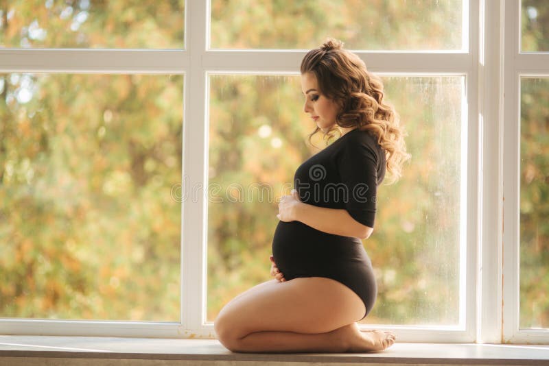 Il moman incinto si siede davanti alla grande finestra Modello vestito in tuta nera Ha messo la sua mano sulla pancia Futuro feli fotografia stock Il moman incinto si siede davanti alla grande finestra Modello vestito in tuta nera Ha messo la sua mano sulla pancia Futuro feli fotografia stock