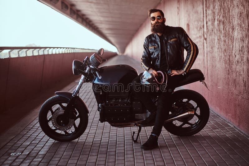Il motociclista alla moda alla moda ha vestito in un bomber nero con le tenute degli occhiali da sole un casco che si siede sul s immagini stock libere da diritti Il motociclista alla moda alla moda ha vestito in un bomber nero con le tenute degli occhiali da sole un casco che si siede sul s immagini stock libere da diritti
