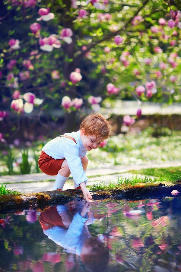 Il piccolo neonato sveglio mette la sua mano nell'acqua dello stagno nel giardino di primavera fotografia stock libera da diritti Il piccolo neonato sveglio mette la sua mano nell'acqua dello stagno nel giardino di primavera fotografia stock libera da diritti