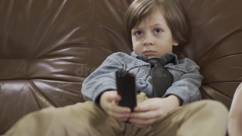 Il piccolo ragazzo sveglio che si siede sul sofà di cuoio con le gambe a parte che cambiano i canali sulla TV facendo uso della r